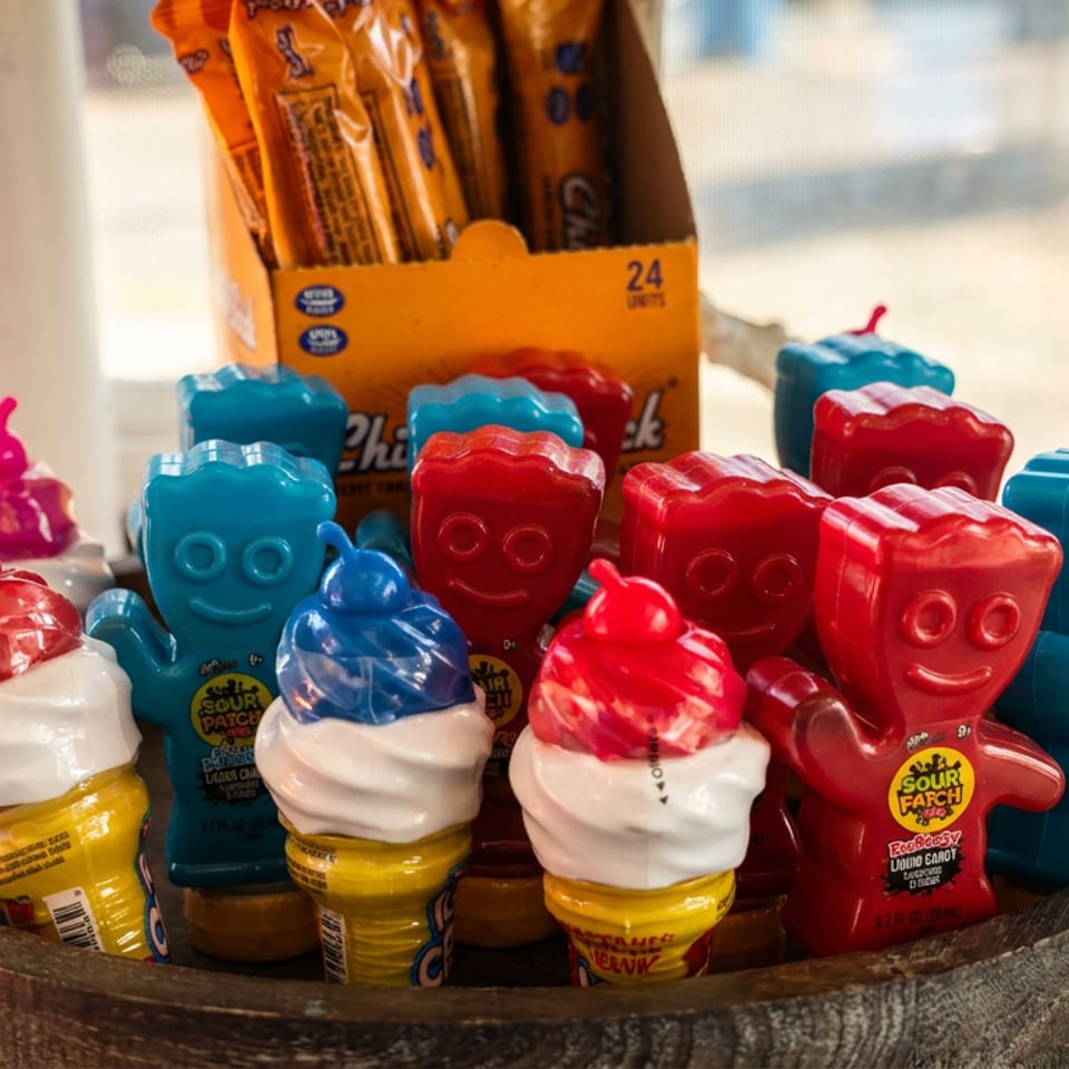 Colorful Sour Patch Kids candy containers and lollipop-shaped treats arranged in a wooden display by a sunlit window.