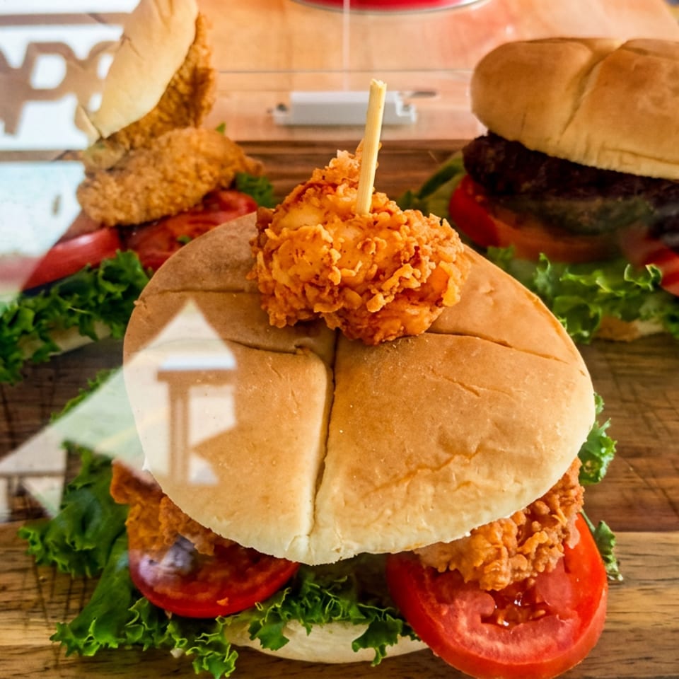 Three burgers with crispy fried chicken, beef patties, lettuce, and tomatoes displayed on a wooden counter.